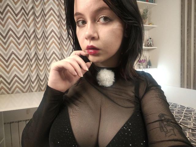 Elfrieda Leever, con cabello negro, lápiz labial rojo y una blusa negra transparente que muestra sus grandes senos, con un dedo cerca de sus labios brillantes, un collar blanco esponjoso y un tatuaje en el brazo derecho..