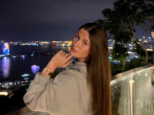 LakeishaCresencio with long brown hair, gray hoodie, and gold watch, looking over shoulder, night city lights in background, hand near mouth, reflective water, trees to the right.
