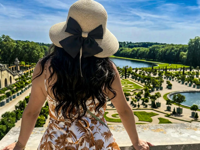 ZoeeMar with long wavy black hair, wearing a sunhat with a black bow, floral dress, and standing with arms on a stone railing, facing a lush garden.