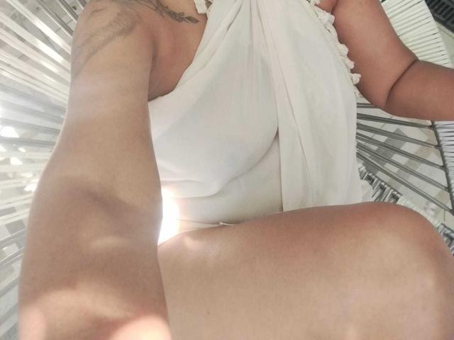 ExoticLylas weißes Kleid, das ihre Brüste und ihren Hintern zeigt, mit ihrem tätowierten Arm, aufgenommen aus einem niedrigen Winkel, wobei ihre gebräunte Haut und das Sonnenlicht auf ihrem Oberschenkel zu sehen sind..