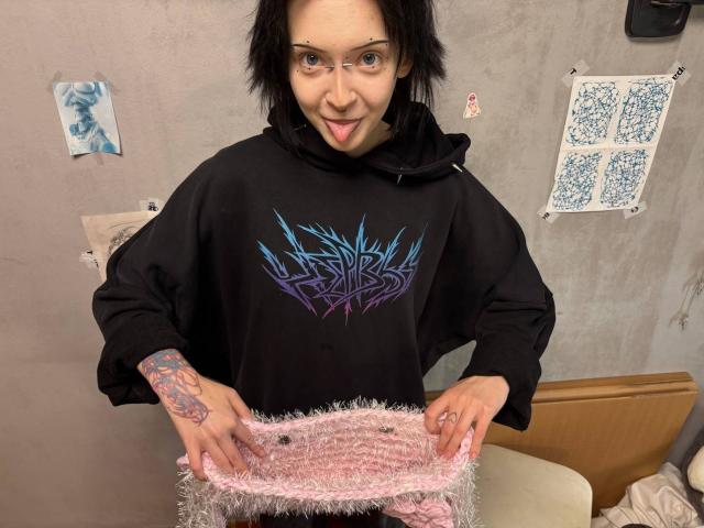 LianeBadenoch with black hoodie, blue-purple graphic, tongue out, holding fluffy pink ass-warmers, tattooed arms, glasses, short black hair, in a room with wall art.