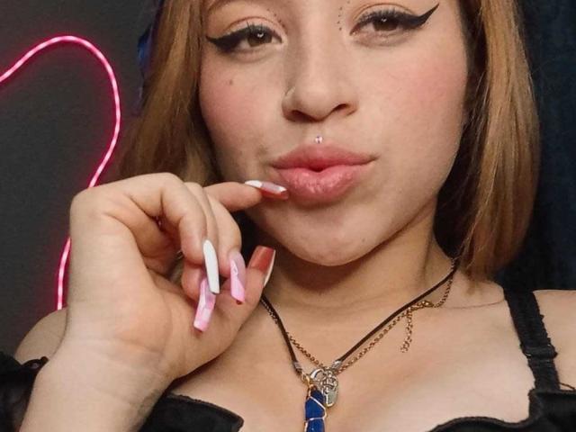 MelodyAnal has a nose ring, lip piercing, and pink manicured nails, wearing a black top with a blue gem necklace, and has a cute, pouty expression.