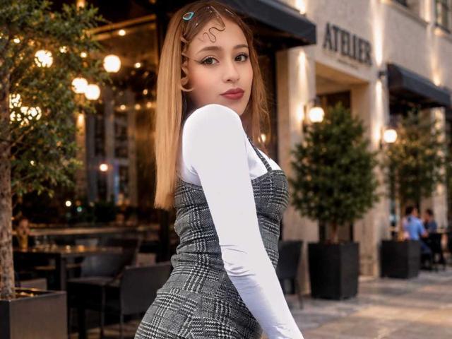 MelodyAnal a les cheveux blonds, porte une chemise blanche à manches longues et une robe grise à carreaux, et se tient dehors avec une jolie pince à cheveux. Elle regarde par-dessus son épaule, montrant ses fesses..