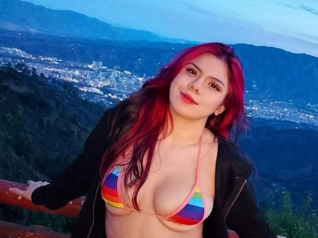 MaraCortes with red hair, wearing a rainbow bikini top, showing her tits, black jacket, smiling, arms on wooden railing, twilight mountain city view in background.