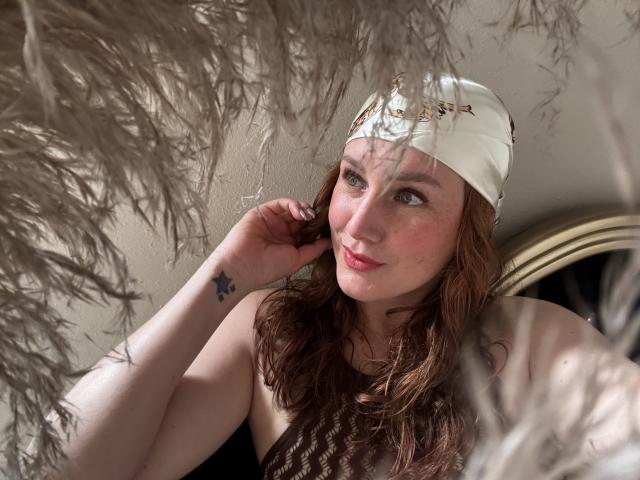 KatyaRogersX with red wavy hair, white cap, freckles, small star tattoo on left wrist, smiling, blue eyes, bare shoulders, brown top, surrounded by fluffy beige feathers.