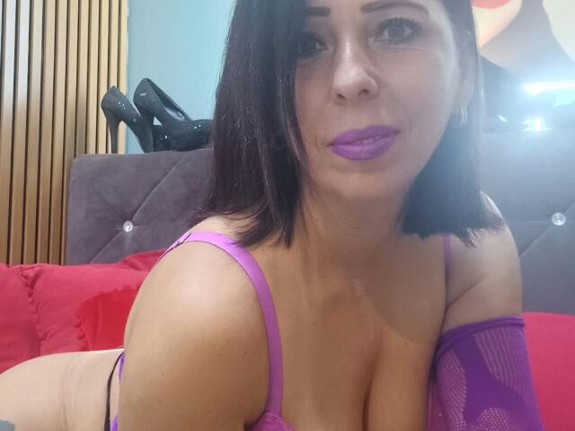 LauraSin with purple lipstick and black hair, wearing a purple bra and arm sleeves, showing her tits, sitting on a red pillow, and black high heels in the background.