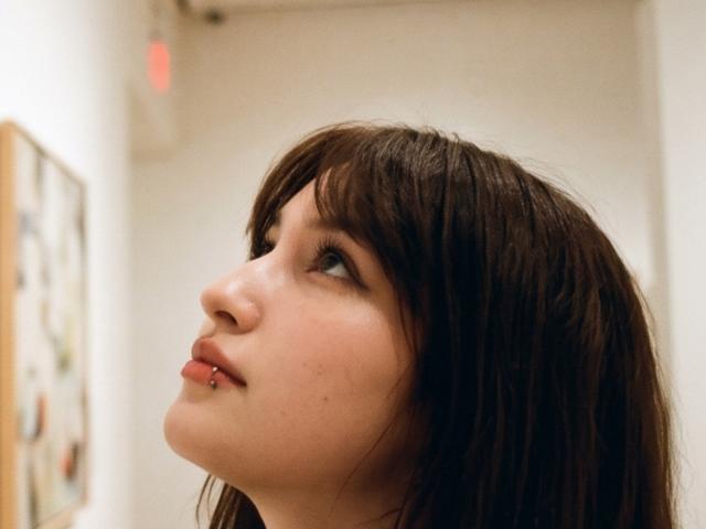 Trisha Ridderhoffs Gesicht im Seitenprofil, braunes Haar, leicht hochgezogene Mundwinkel, kleine Nase, durchstochene Unterlippe, Blick nach oben, weiches Licht, leichte Röte, leicht geöffneter Mund..