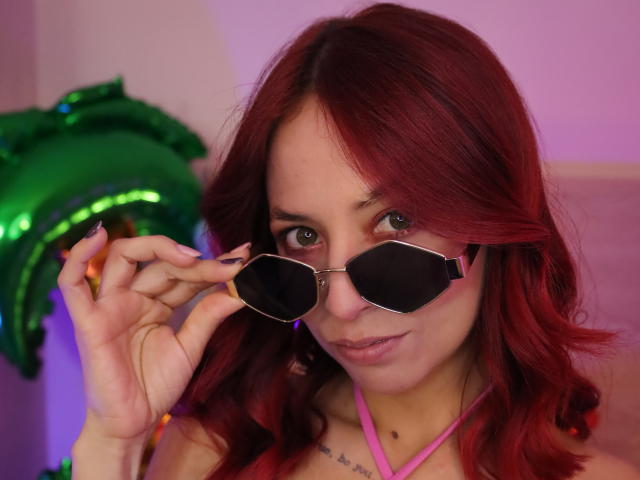 MiaaRouge, con cabello rojo, usando gafas de sol, sosteniéndolas con su mano derecha, top rosa de tirantes, tatuaje discreto en el pecho, un globo verde en el fondo, una ligera sonrisa, iluminación púrpura..