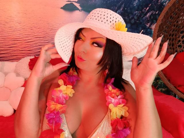 AmberSunshine con un cappello da sole bianco, una ghirlanda di fiori colorati e un trucco giallo sugli occhi, tiene il cappello, mostrando il seno su una sedia imbottita rossa, appare carina..
