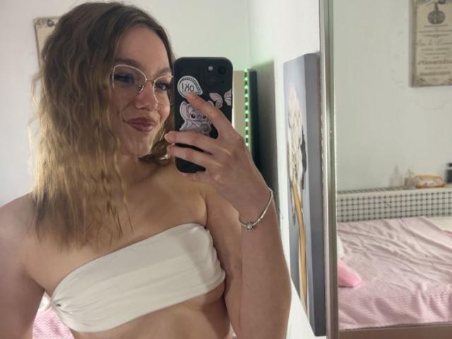LeilaBie, avec ses cheveux blonds ondulés, ses lunettes et un haut blanc sans bretelles, se prend en photo devant un miroir, montrant sa petite poitrine, tout en tenant un téléphone avec un autocollant..