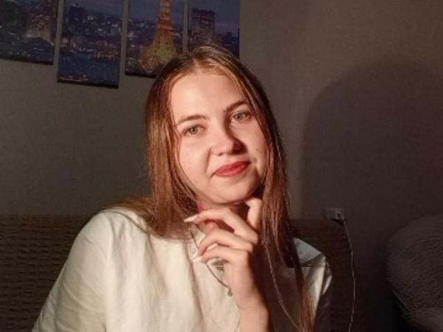 TanyaTandus with wet brown hair, light skin, and red lipstick, wearing a white shirt, touching her chin, in a dimly lit room, showing a subtle smile.