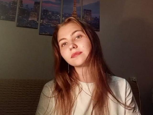TanyaTandus with long brown hair, white shirt, light skin, small moles on her face, slight smile, looking at camera, small nose, subtle makeup, dark room, soft light.