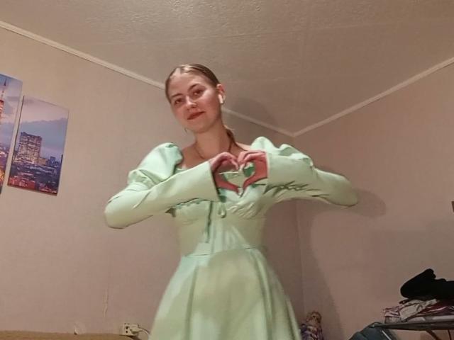 TanyaTandus with blonde hair in a green dress, hands forming a heart, standing in a room, wearing white earbuds, smiling slightly, pale skin, small breasts, cute face.