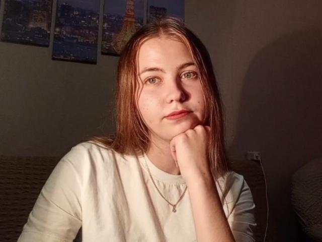 TanyaTandus with light brown hair, green eyes, and a white shirt, resting her chin on her hand, wearing a necklace, in a dimly lit room.