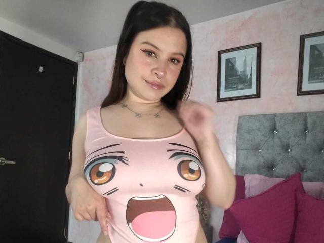 MelaniBecker with long brown hair, wearing a pink anime-style tank top with big eyes and an open mouth, pointing at her big tits, smiling in a bedroom.