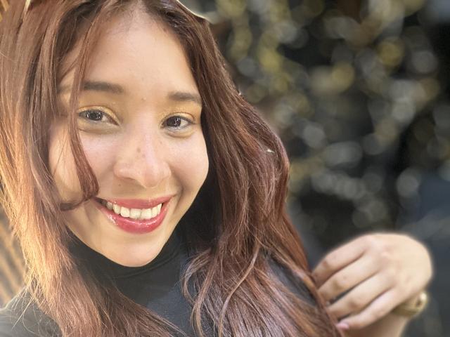 JaneKetty a un sourire charmant, du rouge à lèvres, et des cheveux bruns ondulés, avec une main dans les cheveux, elle porte un pull noir à col roulé, et ses yeux marrons sont fixés sur l'appareil photo..
