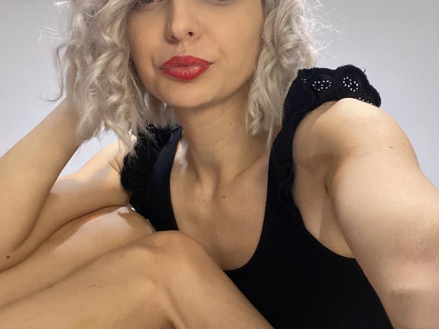 DouceAmelia with curly white hair, red lipstick, black sleeveless top, sitting with knees up, showing her bare arms and tits, looking at camera with a slight smile.