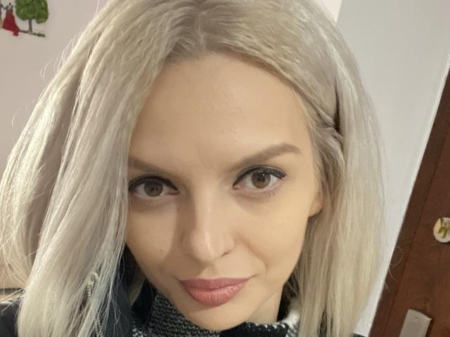 DouceAmelia has pale skin, blonde hair, and subtle makeup with pink lipstick. She has brown eyes and a slight smile. Her tits are not visible.