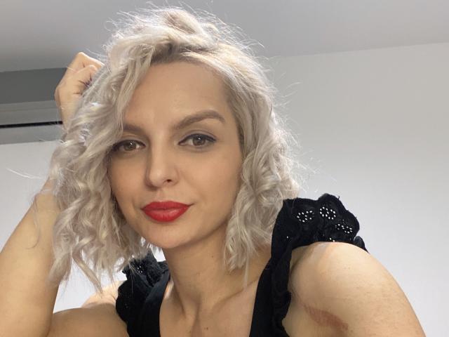 DouceAmelia has curly white hair, red lipstick, and black lace top, looking at camera with hand in hair, showing off her tits and cute face.