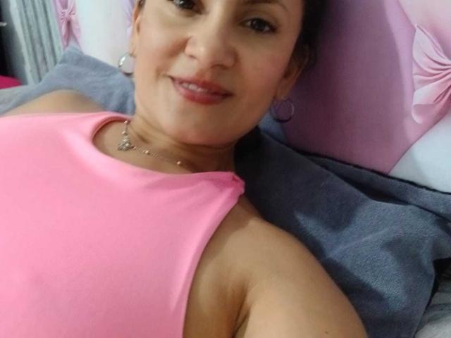SolaraDubois with small tits, pink tank top, smiling, lying down, gray pillow, pink earbuds, necklace, brown hair, light skin, soft light, relaxed, confident, cute.