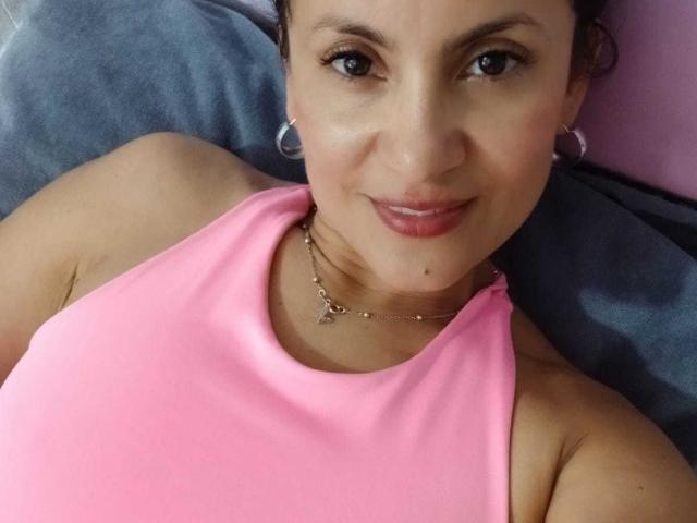 SolaraDubois with brown eyes, pink tank top, hoop earrings, necklace, and subtle smile, lying down, showing her tits and small mole above her lip.