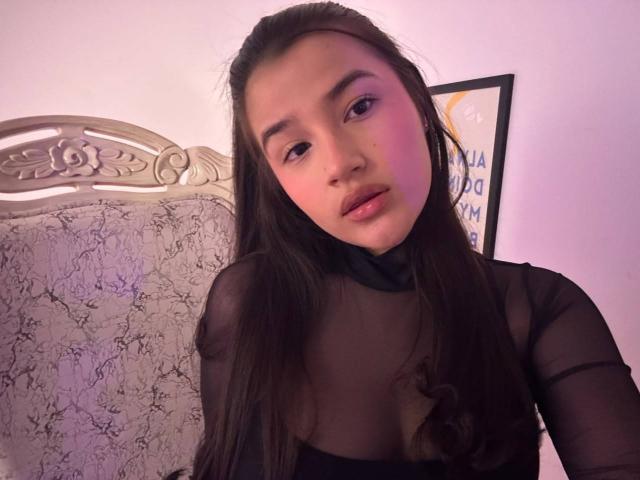 IrisBlair with long brown hair, wearing a sheer black top, looking at the camera with pinkish-purple lighting on her face, showing her small tits and subtle makeup.