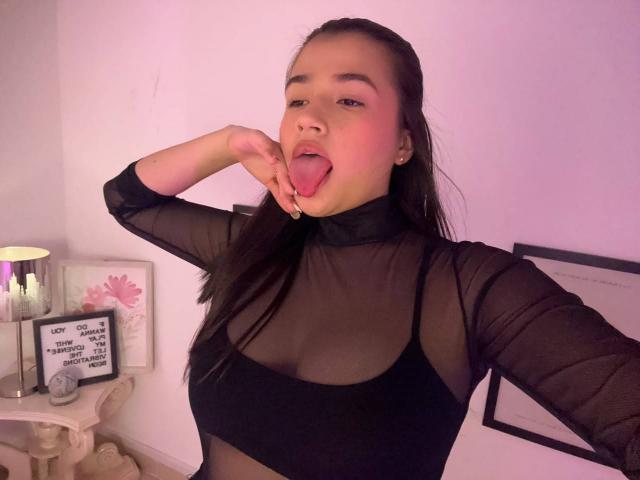 IrisBlair with long dark hair, sticking her tongue out, wearing a sheer black top that reveals her tits, and a nose ring, in a pink-lit room.