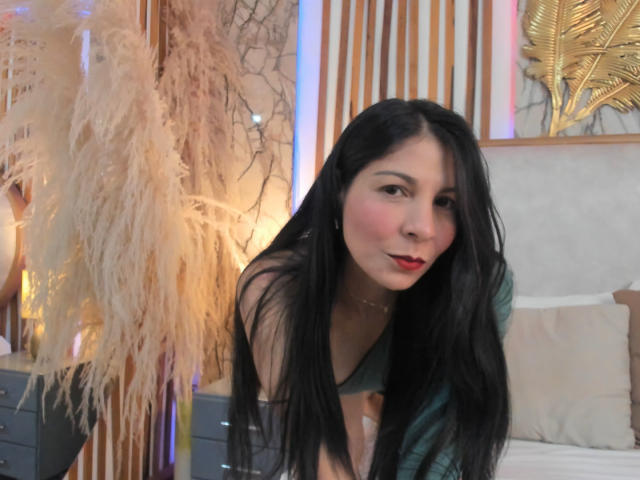 EliseBell with long black hair, red lipstick, and green top, leaning forward, showing her tits slightly, in a softly lit bedroom with pampas grass and gold decor.