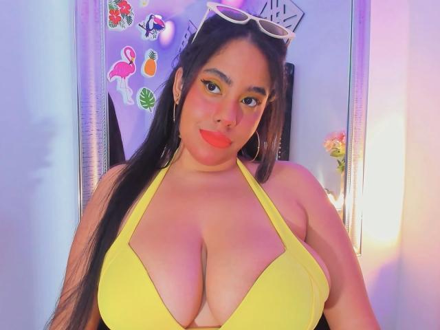 Sienna Taylor, avec un haut de maillot de bain jaune et ample qui met en valeur sa poitrine généreuse, de longs cheveux noirs, du rouge à lèvres rouge, du fard à paupières jaune et des lunettes de soleil blanches sur la tête, souriant à l'appareil photo..