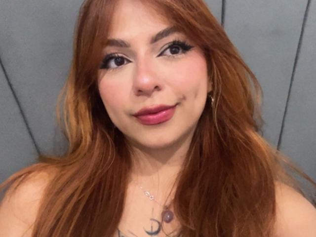 SamantaCarterr has long, reddish-brown hair, thick eyebrows, black eyeliner, pink lipstick, and a small smile. She's wearing gold hoop earrings and a necklace with a purple gem.