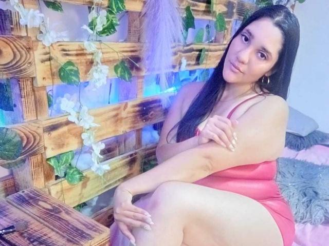 CristalRios with long black hair, pink dress, sitting on a wooden bench, legs crossed, arms folded, cute smile, blue light, green leaves, and white flowers in the background.