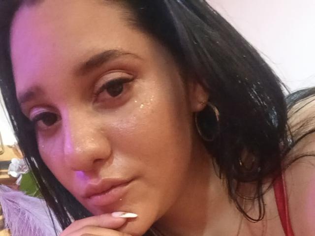 CristalRios has shiny, brown skin with a slight sheen, dark straight hair, and is biting her index finger with white nails. She has hoop earrings and a red top.