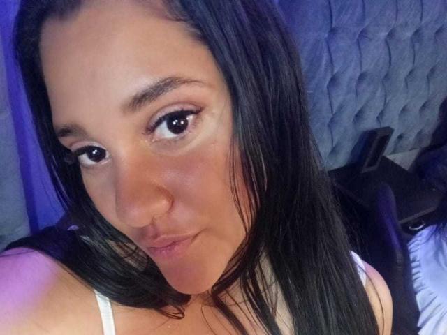 CristalRios has long black hair, brown eyes, and a slightly flushed face. She's wearing a white top, looking up with a subtle pout. Her tits are partially visible.
