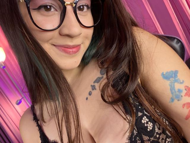 EmmaFeri with long brown hair, glasses, and a black lace top, showing tattoos on her chest and arm, smiling at the camera, with pink and purple lighting. Boobs.