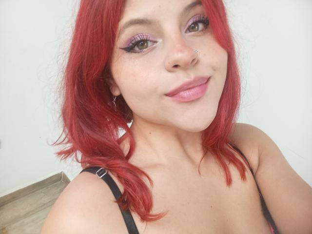 Sophie Hill, avec ses cheveux roux, son eye-liner violet et son rouge à lèvres rose, porte un haut noir à bretelles, a un piercing au nez et affiche un léger sourire, dévoilant sa poitrine..
