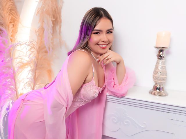AngelRoussy with pink lingerie, lace bra, and sheer robe, smiling, leaning on white table, hand on chin, purple and pink hair, silver candle, white wall, and feather decor.