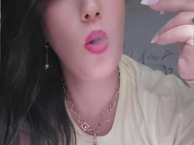 VanessaSquirtt's face, pink lipstick, slightly open mouth, long black hair, gold necklace, pale skin, yellow shirt, manicured nails, right hand near face, soft light, slight shadow.