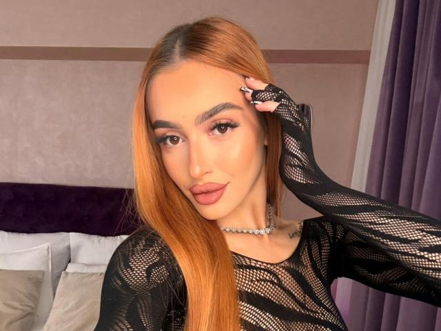 MarissaFex mit langen, orangefarbenen Haaren, einem schwarzen Netzoberteil und rosafarbenen Lippen, die sich mit einer Hand das Haupt berührt, trägt eine Diamantkette, zeigt ihre Brüste und ein Tattoo auf ihrer Brust..