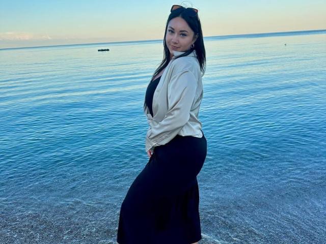 AdriannaGoldis with long black hair, wearing a beige shirt and black skirt, standing by the water, smiling at the camera, sunglasses on head, blue water, pebbled shoreline.