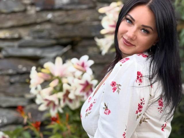 AdriannaGoldis with black hair, blue eyes, and a smile, wearing a white floral dress, showing her tits, against a stone wall with pink and white flowers.
