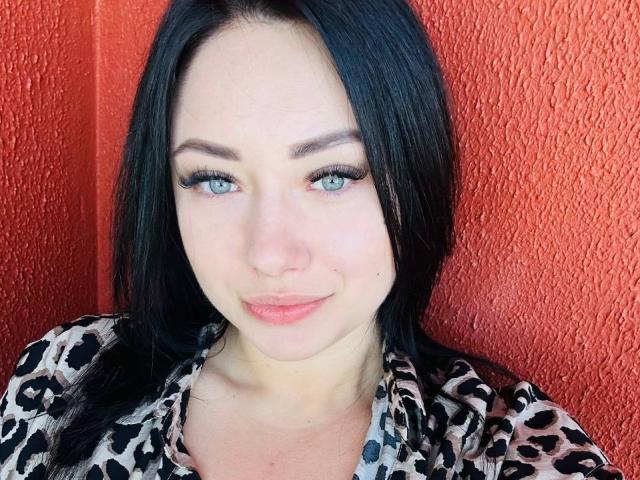 AdriannaGoldis has blue eyes, black hair, and a cute smile, wearing a leopard print shirt. Her tits are not visible, and she's against a textured red wall.