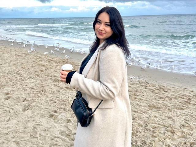 AdriannaGoldis with dark hair, beige coat, black bag, holding coffee, standing on sandy beach, seagulls in background, overcast sky, smiling, tits covered, curvy, cute.