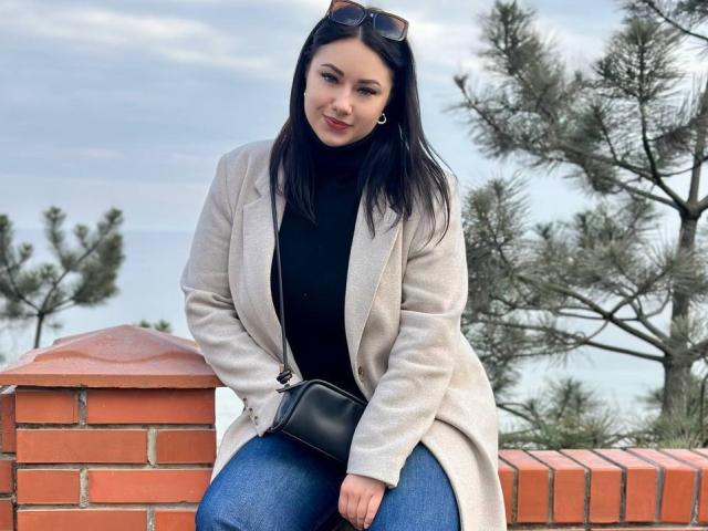 AdriannaGoldis with long black hair, wearing a beige coat, black turtleneck, and blue jeans, sitting on a brick wall, holding a black purse, with sunglasses on her head.