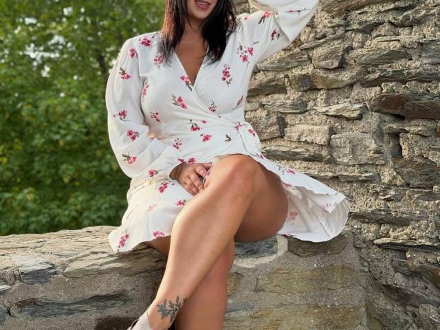 AdriannaGoldis, wearing a white floral dress, sits on stone, showing her bare legs and a small tattoo on her right ankle, with her arm raised, smiling.