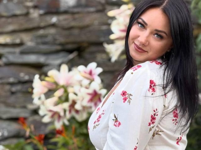 AdriannaGoldis with black hair, blue eyes, and a smile, wearing a white floral shirt, showing her tits, standing in front of pink and white lilies.