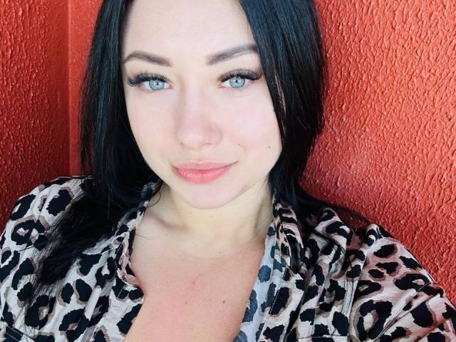 AdriannaGoldis with blue eyes, black hair, and a leopard print shirt, smiling slightly. Her tits are partially visible. Red textured wall in background.