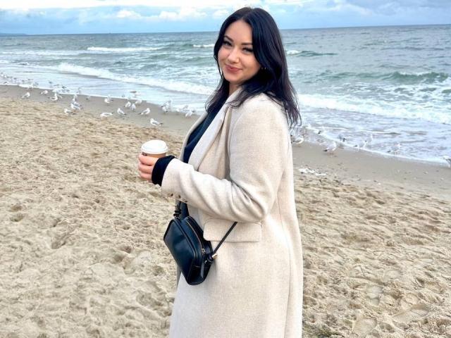 AdriannaGoldis with dark hair, wearing a beige coat, black purse, and holding a coffee cup, standing on a sandy beach with seagulls in the background.