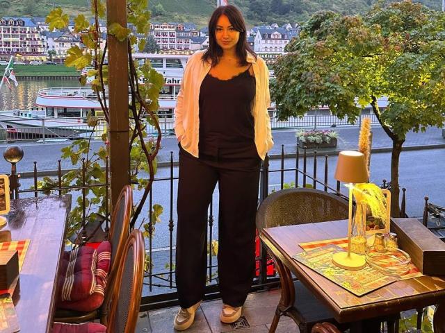 AdriannaGoldis with dark hair, black pants, and white shirt standing on a wooden terrace, hands in pockets, looking forward, green shoes, table with lamp and yellow placemat, leafy tree.