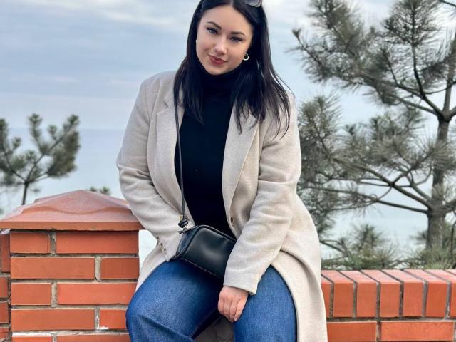 AdriannaGoldis with long black hair, wearing a beige coat, black turtleneck, and blue jeans, sitting on a brick wall, holding a black clutch, and smiling.