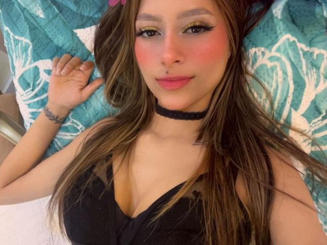 AfroditaWild with long brown hair, black choker, yellow eyeshadow, pink blush, black top showing her big tits, lying on a teal floral pillow, smiling at the camera.
